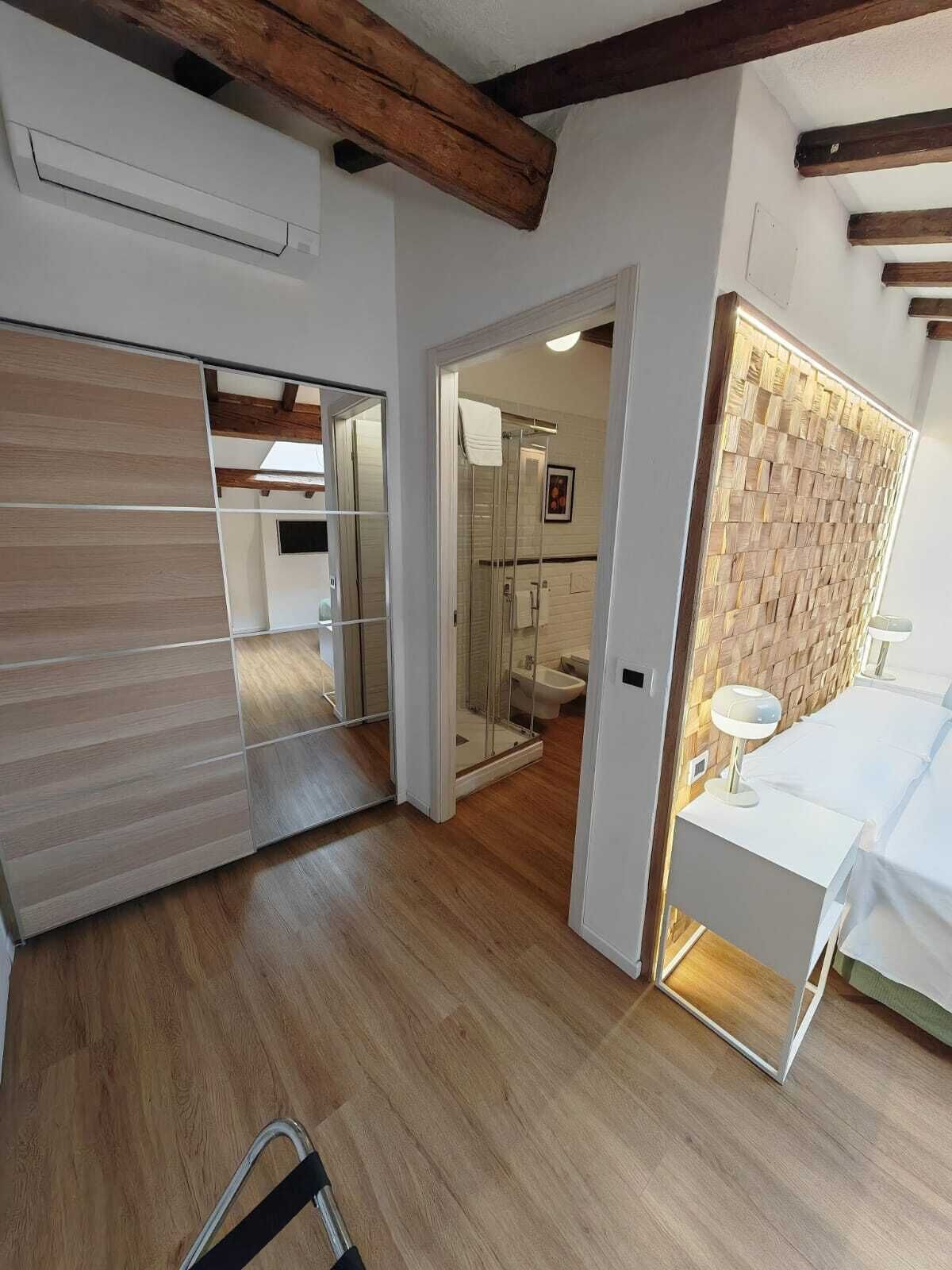 Ein modernes Hotelzimmer mit Holzboden, einem großen Spiegel, einem begehbaren Kleiderschrank, einer Duschkabine und einem Nachttisch mit Lampe.