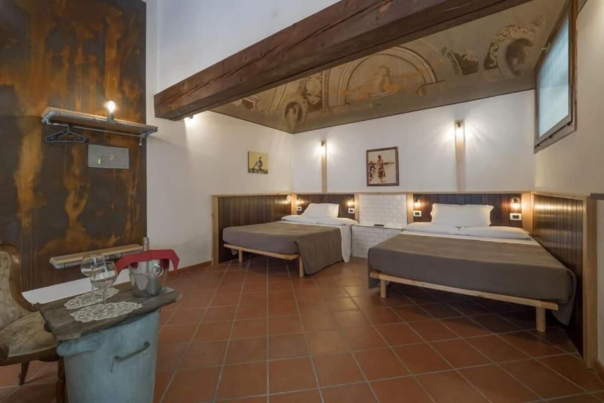 A room with two beds, a wooden bench, a small table with a wine glass, and a wall with a mural.