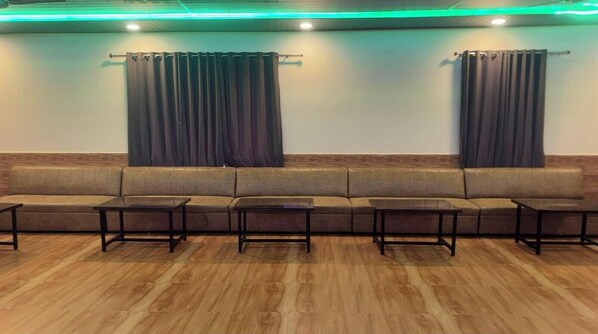 A waiting area with a long sofa and small tables, wooden flooring, and curtains on the windows. - The Peace In Mountain Hotel (Kotdwāra)