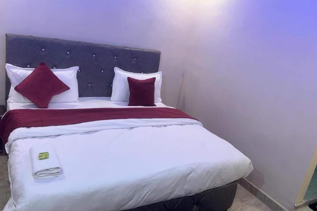A neatly made bed with white and red bedding, a grey tufted headboard, and a small towel on the footboard.