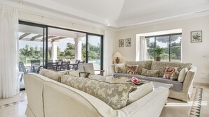 Amplia sala de estar con un sofá grande, comedor con sillas y una mesa, y ventanas grandes que ofrecen vista a zonas verdes.
