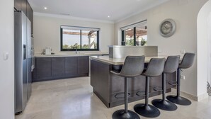 Una cocina moderna con armarios oscuros, una isla central con taburetes de bar, un ventanal grande y un reloj fijado a la pared.