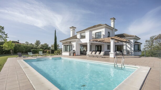 Una piscina rectangular con solárium alicatado, un edificio blanco con tejado de baldosas y un cielo despejado.