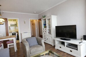 Ein Wohnzimmer mit Fernseher, einer Couch, einem Esstisch und einer Küche im Hintergrund.