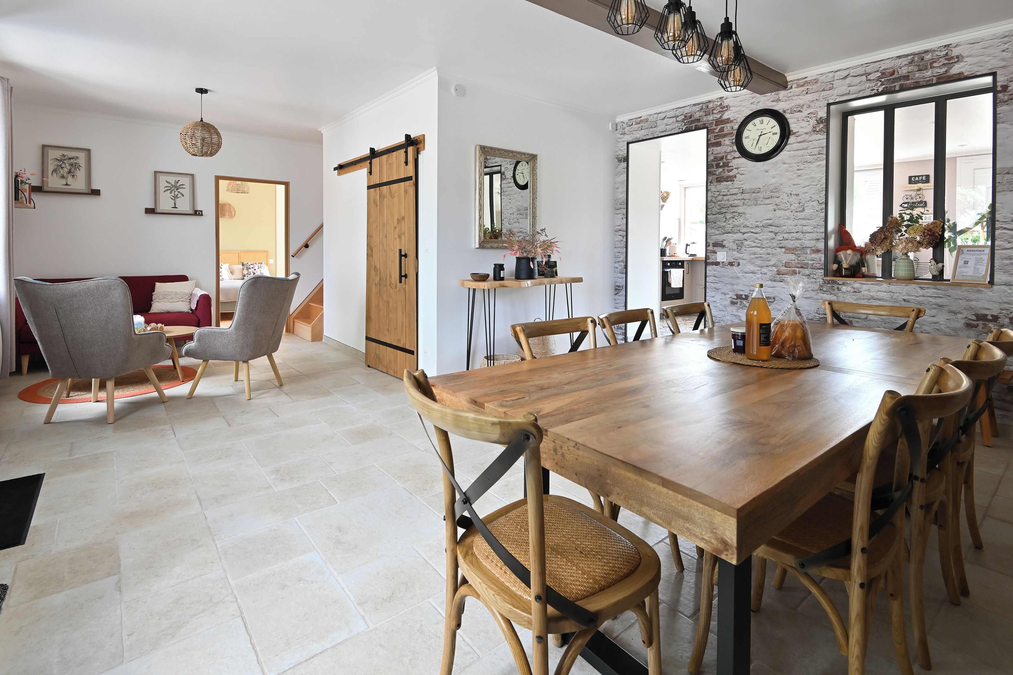 Un comedor espacioso con una larga mesa de madera y varias sillas, una zona de cocina con pared de ladrillo, y una sala de estar con un sofá rojo y un sillón gris.