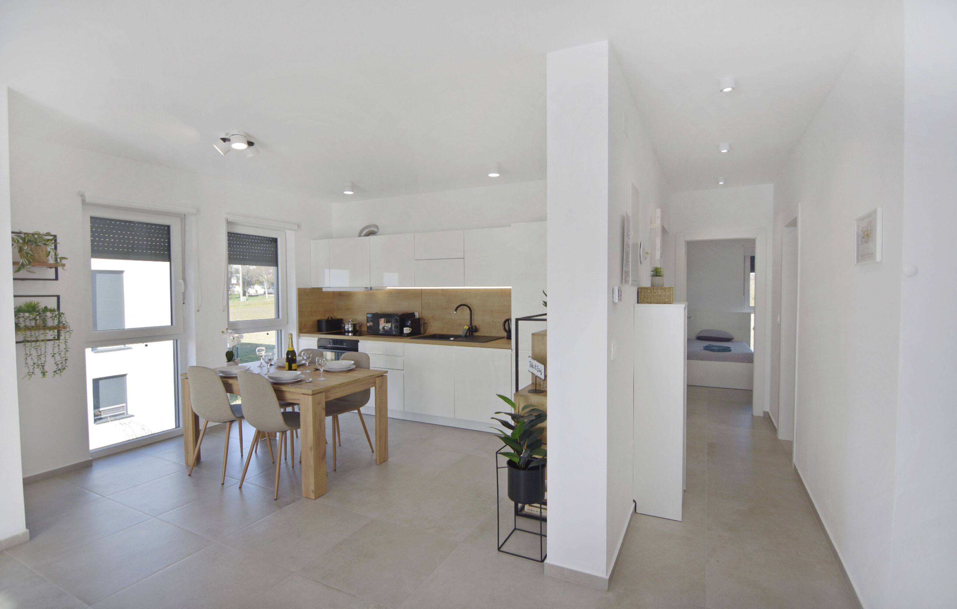 Une cuisine moderne avec des meubles blancs, une table à manger en bois et des chaises. On y trouve un petit îlot central avec un évier et une plante en pot.