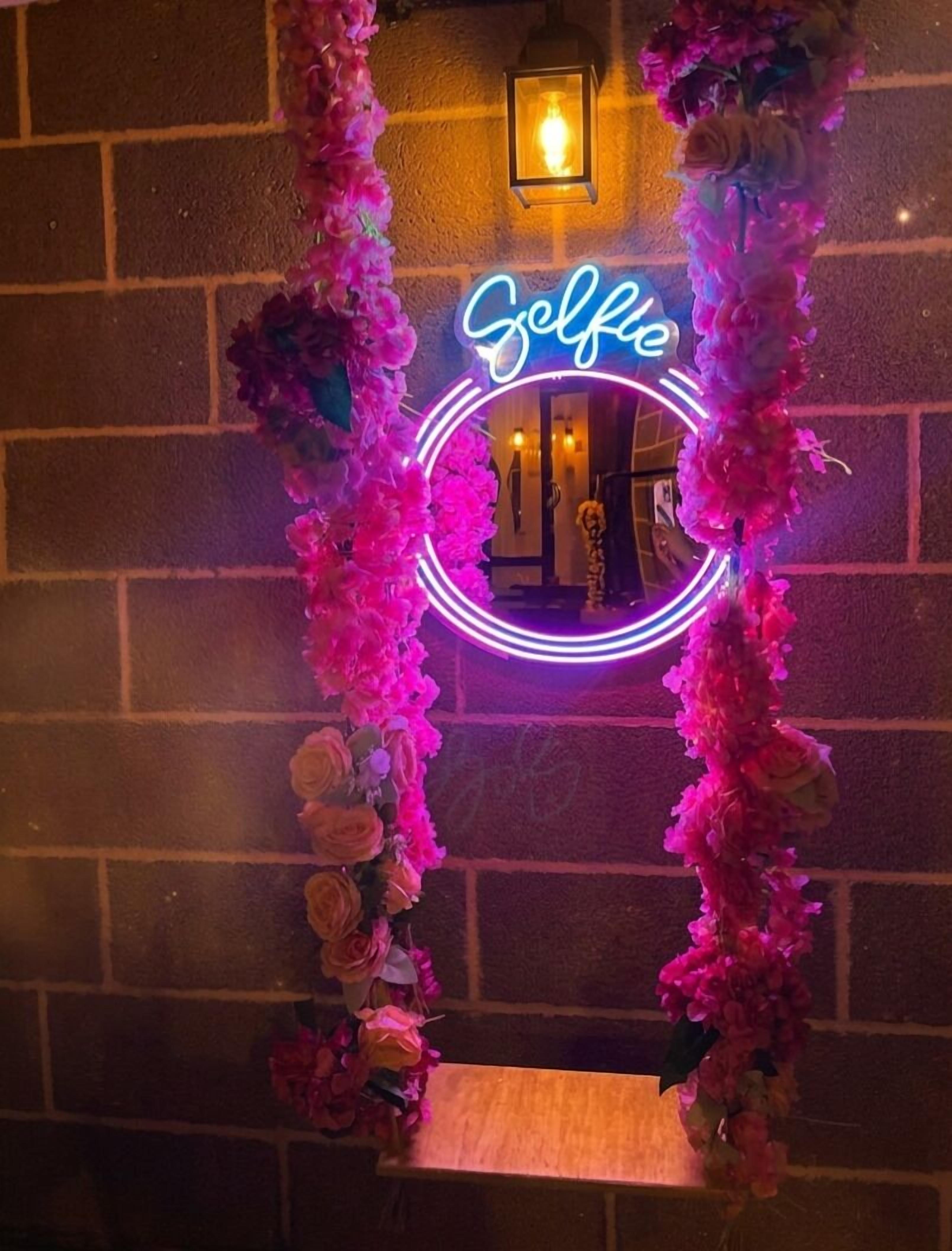 Un letrero de neón con la palabra 'Selfie' en un marco circular, rodeado de decoraciones florales rosas sobre una pared de ladrillo.