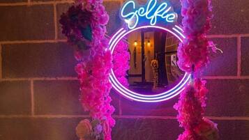Un letrero de neón con la palabra 'Selfie' en un marco circular, rodeado de decoraciones florales rosas sobre una pared de ladrillo.