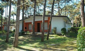 A single-story house with a red and white exterior, surrounded by trees and a grassy yard. - Villa with patio in Lignano Pineta (Lignano Sabbiadoro)