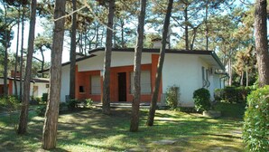 A single-story house with a red and white exterior, surrounded by trees and a grassy yard. - Semi-detached house in Lignano Pineta (Lignano Sabbiadoro)