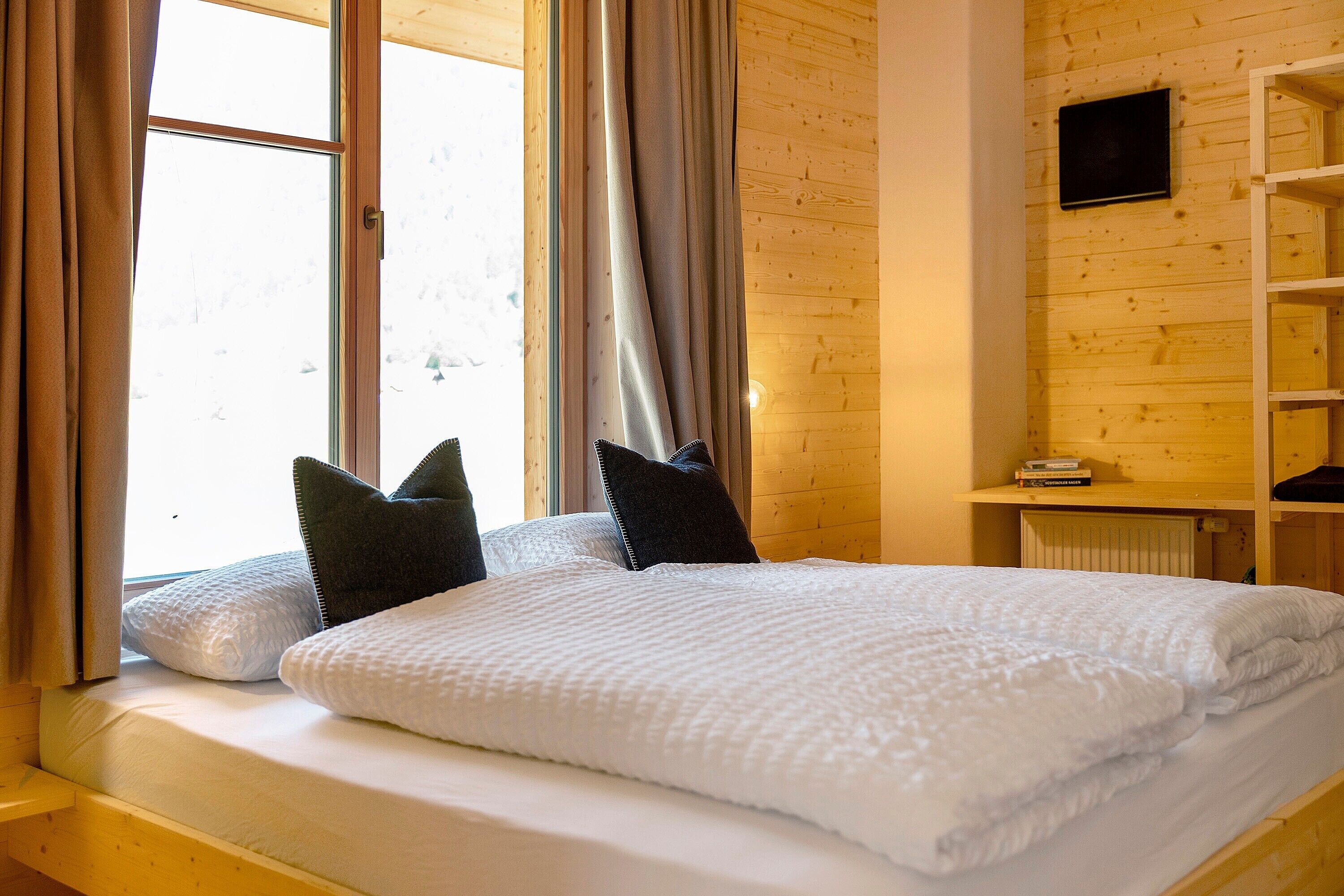 Un lit avec une literie blanche, une fenêtre avec des rideaux et un mur en bois avec une étagère.