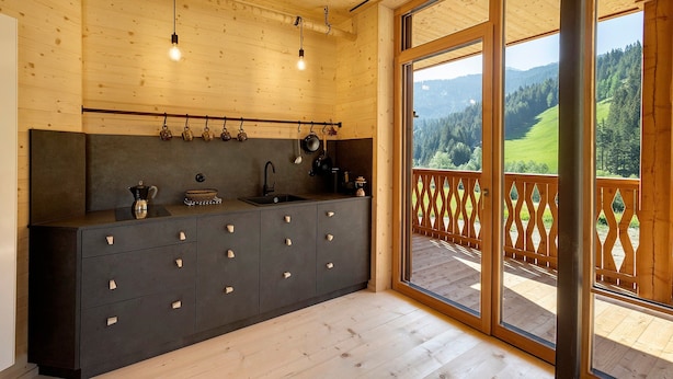 Interior de madera con cocina, un amplio ventanal corredero a una terraza exterior y vistas de un paisaje verde.
