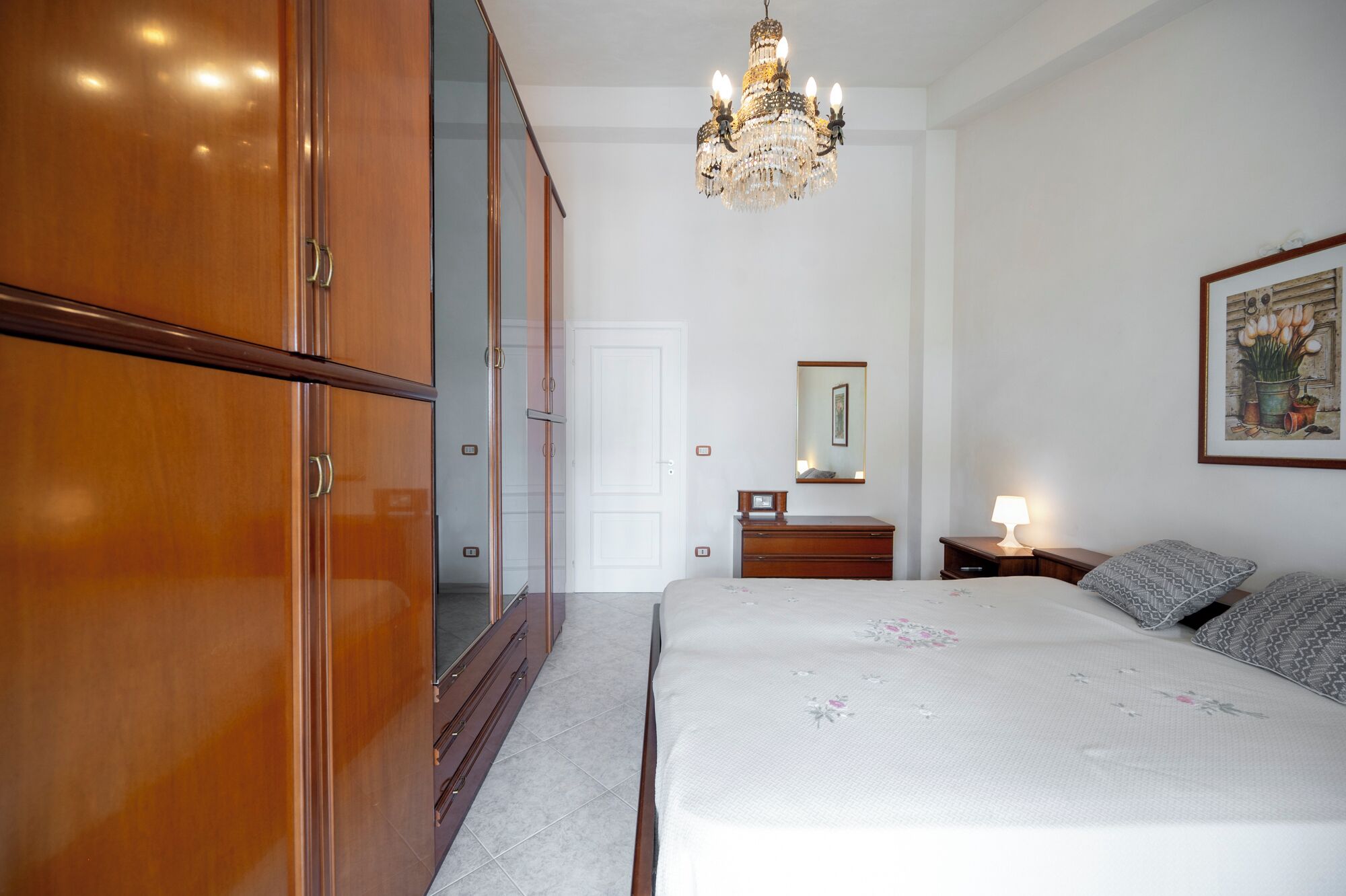 Quarto com guarda-roupa de madeira, cama com colcha florida, mesa de cabeceira com abajur e candelabro.