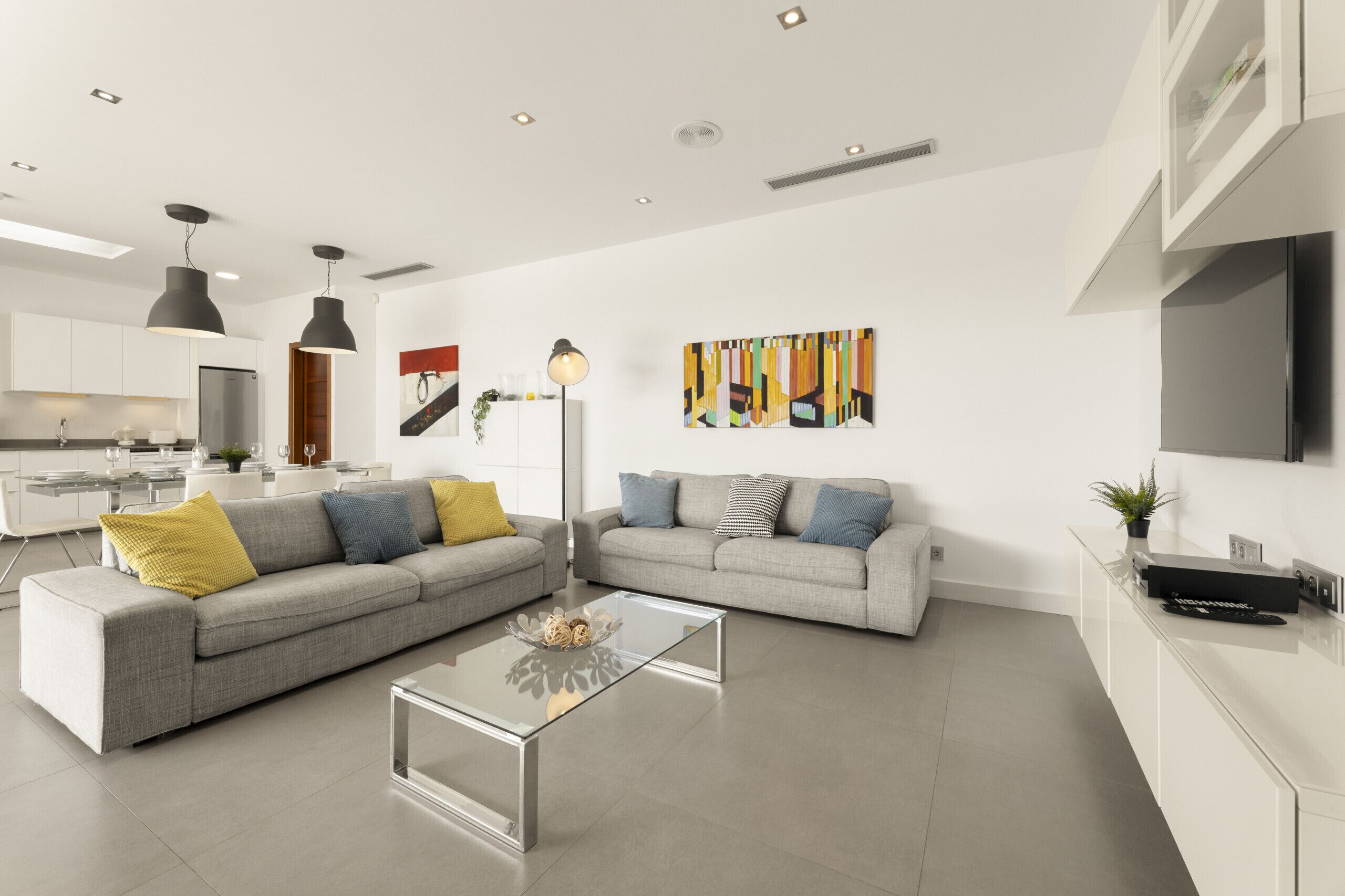 A modern living room with a grey sectional sofa, a glass coffee table, and a minimalist kitchen in the background.