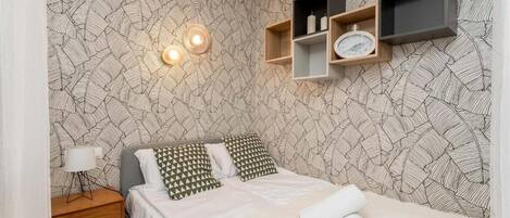 A neatly arranged bedroom with a patterned wallpaper, a bedside table with a lamp, a bed with pillows, and wall-mounted shelves with decorative items.