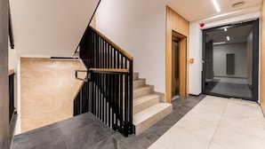 A modern staircase with a wooden handrail and black balusters, leading to a room with a glass door and a wooden panel wall. - Malila Apartments Plac Bohaterów Getta (Kraków)