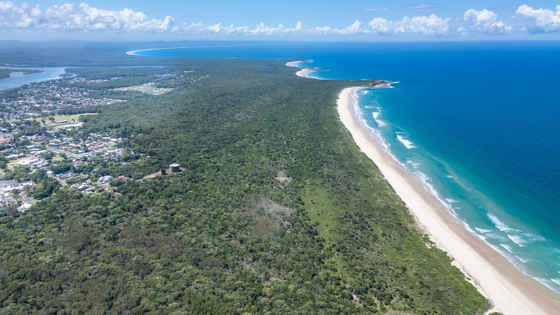 An aerial view of a coastal area with a beach, dense forest, and a small town.