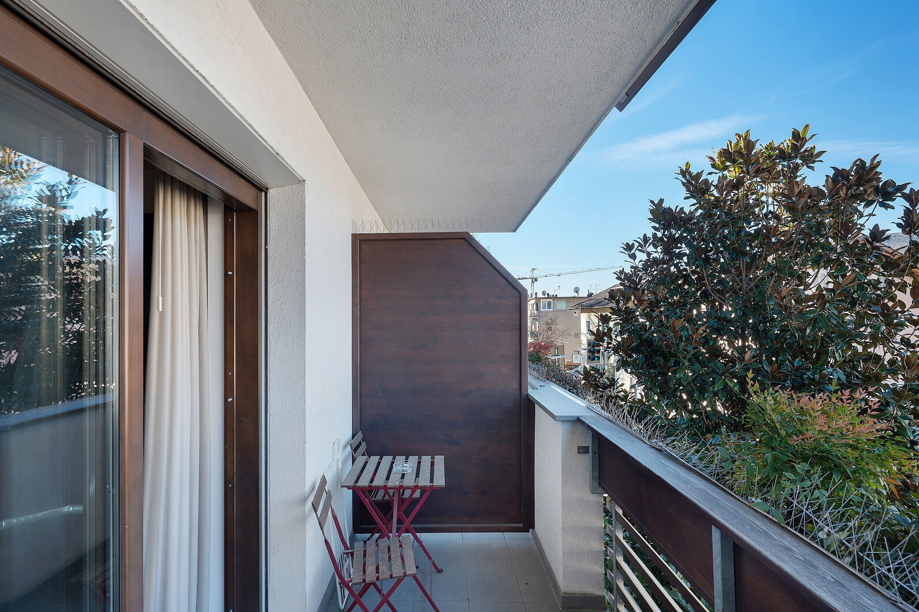 Un balcó amb taula i cadires de fusta, una finestra amb cortines, i vista a un arbre i edificis.