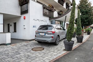 A modern building with the name "ALPENROSE" on the facade, a parked car, and potted plants along the sidewalk.