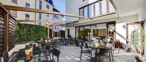 An outdoor patio area with multiple tables and chairs, a glass-enclosed room, and a view of adjacent buildings.