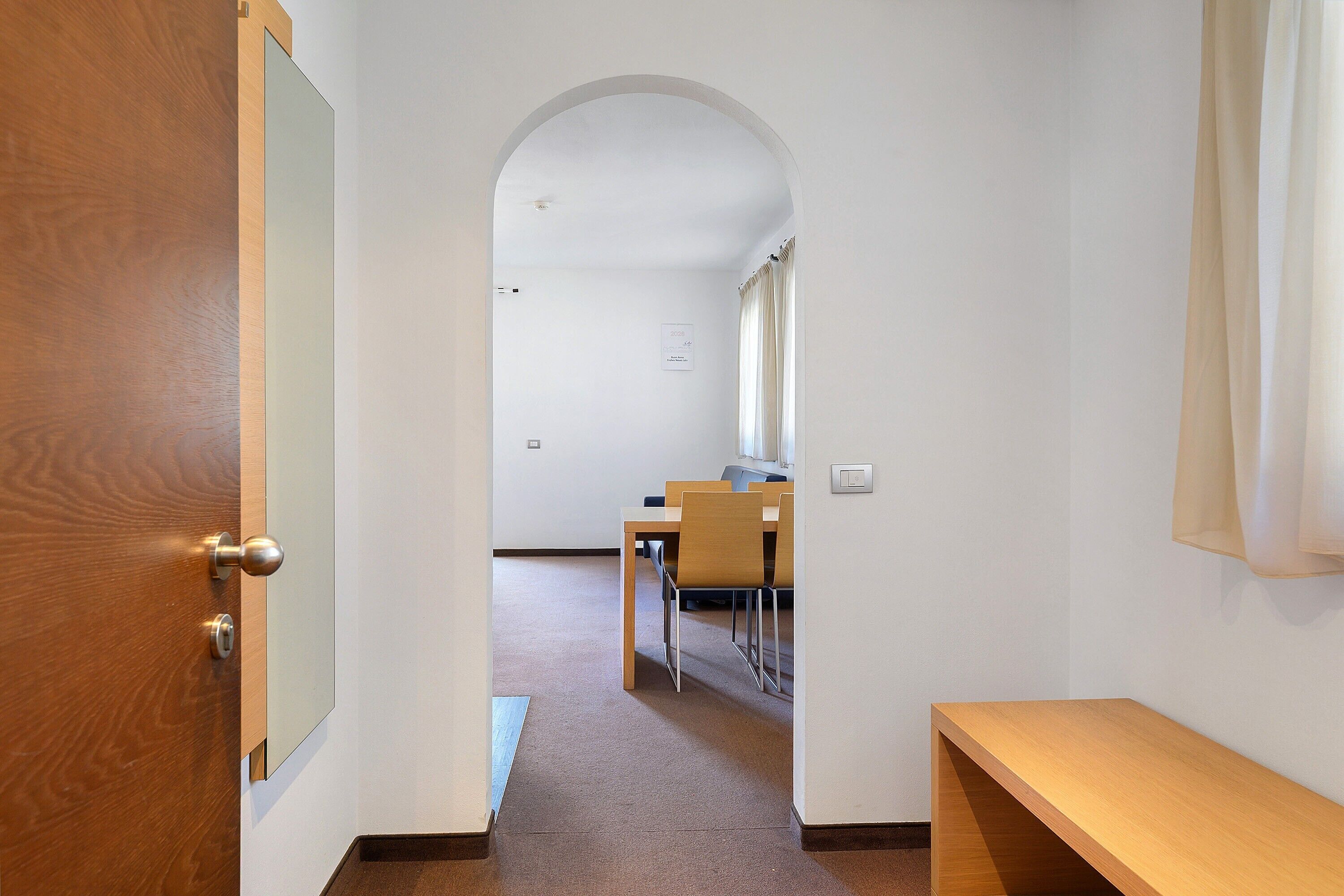 Un pasillo con una puerta de madera, un banco y un comedor con sillas y una mesa.