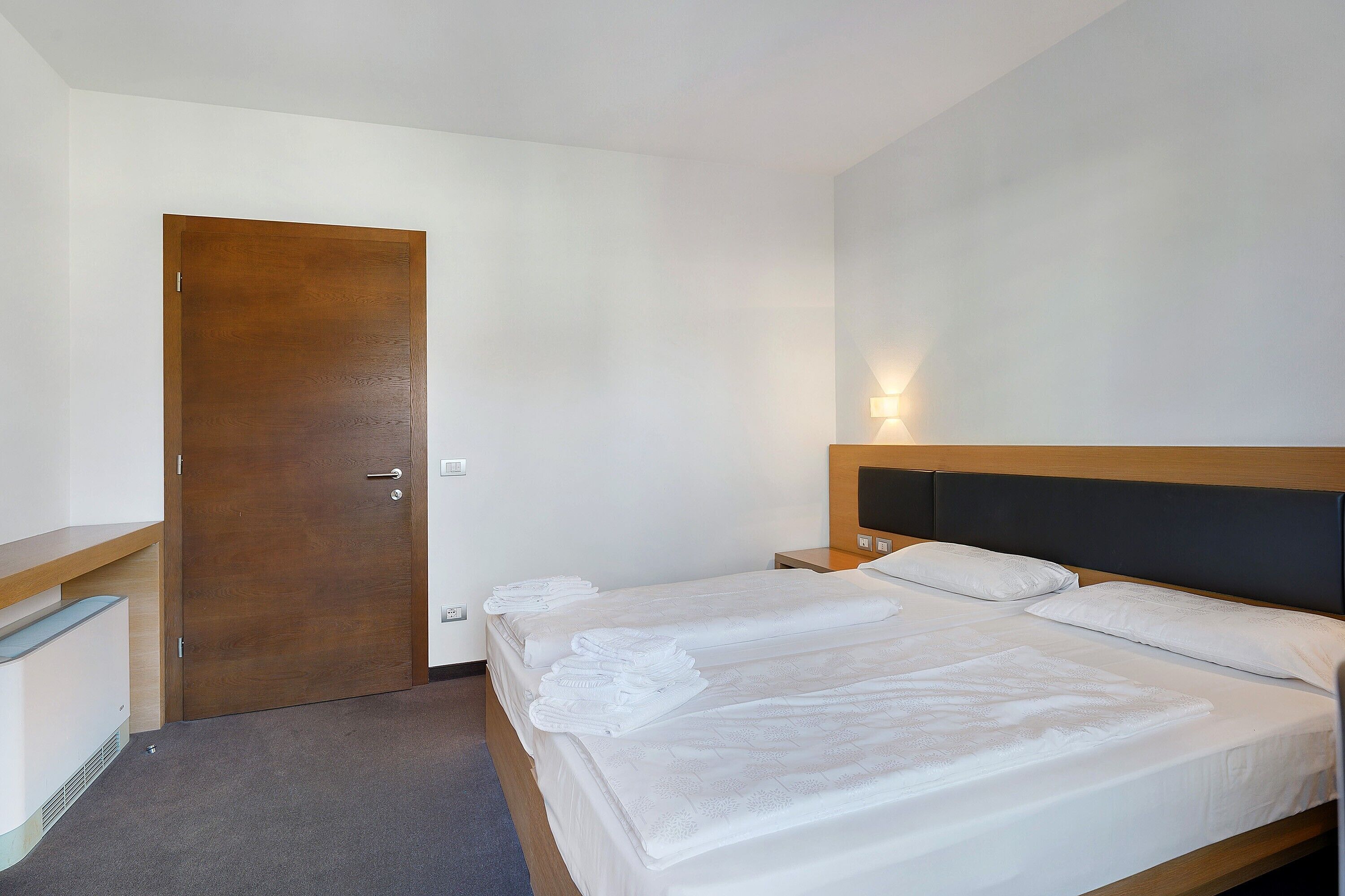 Quarto de hotel com porta de madeira, cama com roupa de cama branca, mesa de cabeceira e luminária fixada na parede.