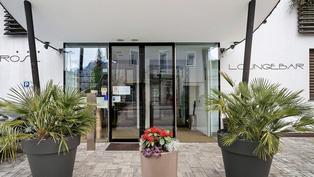 L’entrée d’un immeuble est ornée d’une porte en verre, d’une enseigne indiquant « LOUNGEBAR », et de deux grands plants en pot placés de part et d’autre.