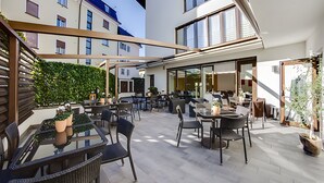 Un espace extérieur aménagé en terrasse, avec plusieurs tables et chaises, une pièce fermée par des baies vitrées et une vue sur les bâtiments adjacents.