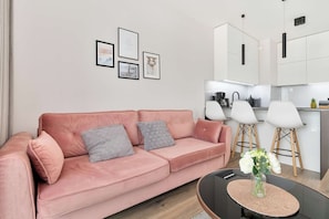 Una sala de estar moderna con un sofá rosa, una mesa de centro y una zona de cocina con armarios blancos y luces colgantes.