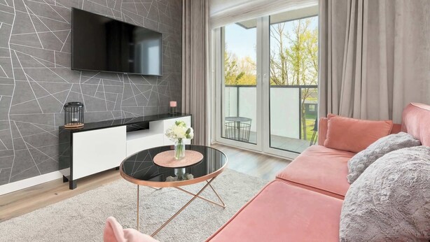 A modern living room with a flat-screen TV mounted on a textured wall, a sofa, a coffee table with a vase, and a balcony with curtains.