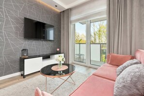 A modern living room with a flat-screen TV mounted on a textured wall, a sofa, a coffee table with a vase, and a balcony with curtains. - Modern Apartment for 4 persons | Parking in the garage (Gdańsk)
