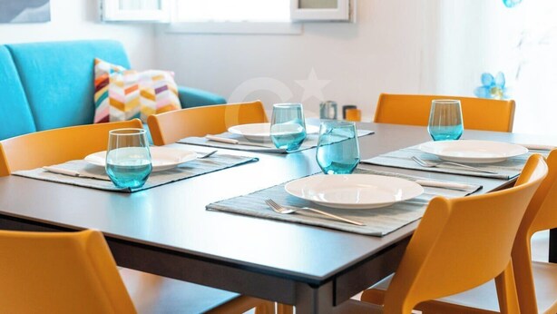 Una mesa de comedor dispuesta con vasos azules, platos blancos y sillas amarillas en una habitación luminosa con un sofá azul y almohadones coloridos.