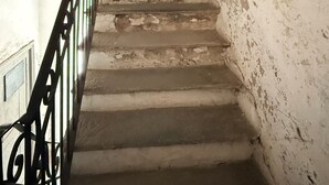 Uma escadaria com degraus desgastados e corrimão de metal, que leva a uma porta escura.