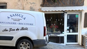 Uma carrinha branca estacionada em frente a uma padaria chamada 'A SASSULA', com uma placa de identificação acima da entrada.