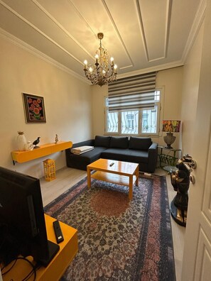 A living room with a black sofa, a wooden coffee table, a TV on a yellow stand, a chandelier, a window with blinds, and a decorative bird statue. - Bohemian Antique House in Colorful Balat (İstanbul)