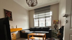 A living room with a sofa, a coffee table, a television, a chandelier, and a window with blinds. - Bohemian Antique House in Colorful Balat (İstanbul)