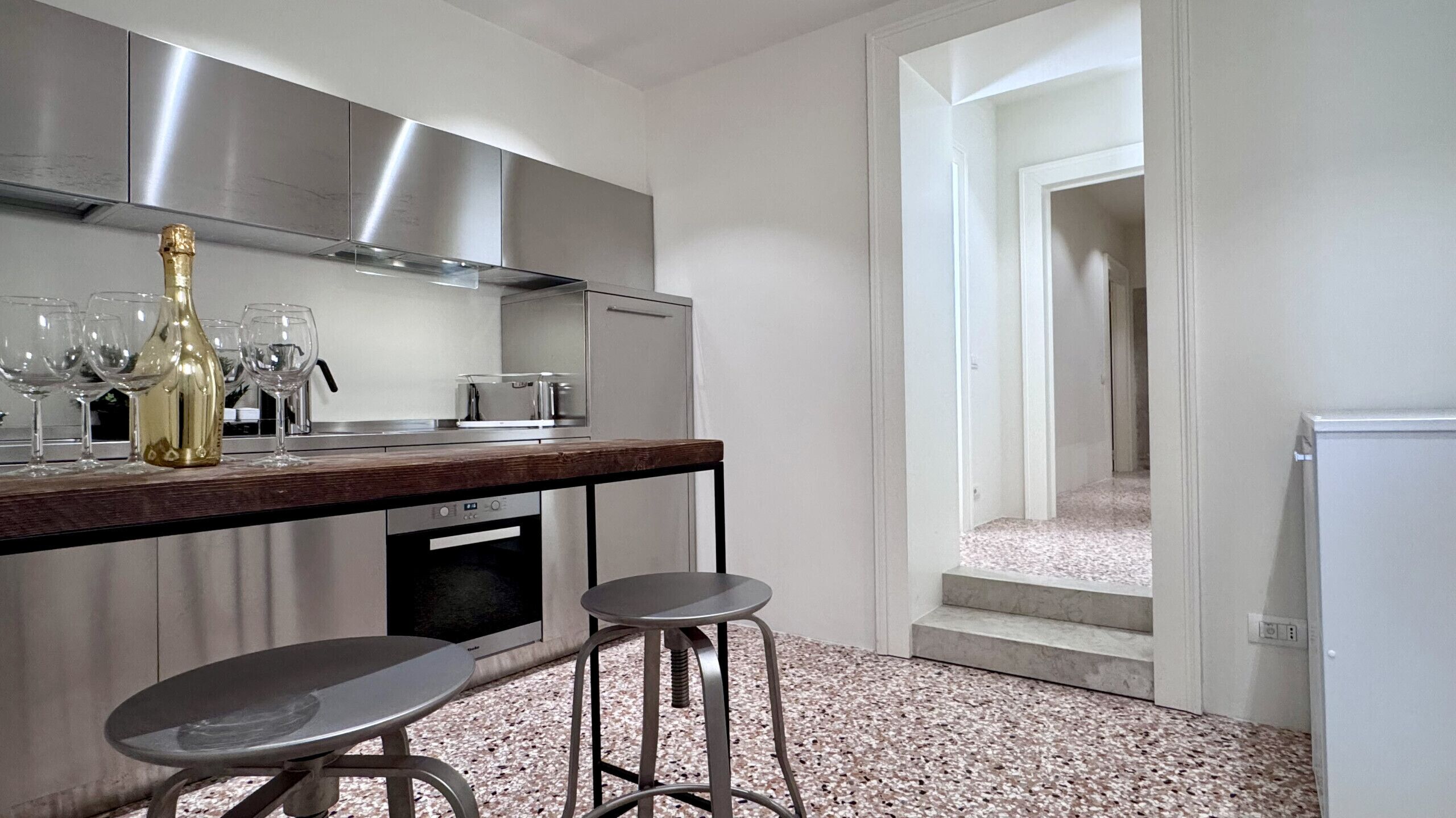 A modern kitchen with stainless steel appliances, a wooden countertop, two bar stools, and a wine bottle.