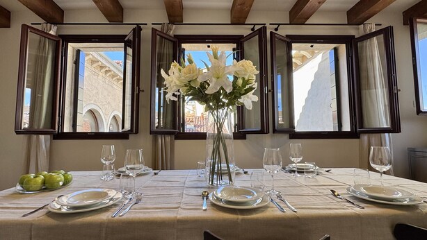 A dining table set for four with a vase of white flowers in the center, surrounded by windows with open shutters.