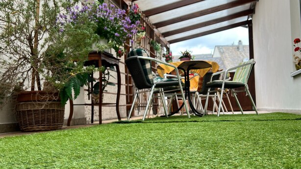 Ein Außenbereich mit Terrasse, Tisch und Stühlen, umgeben von Topfpflanzen und einem Rasen.
