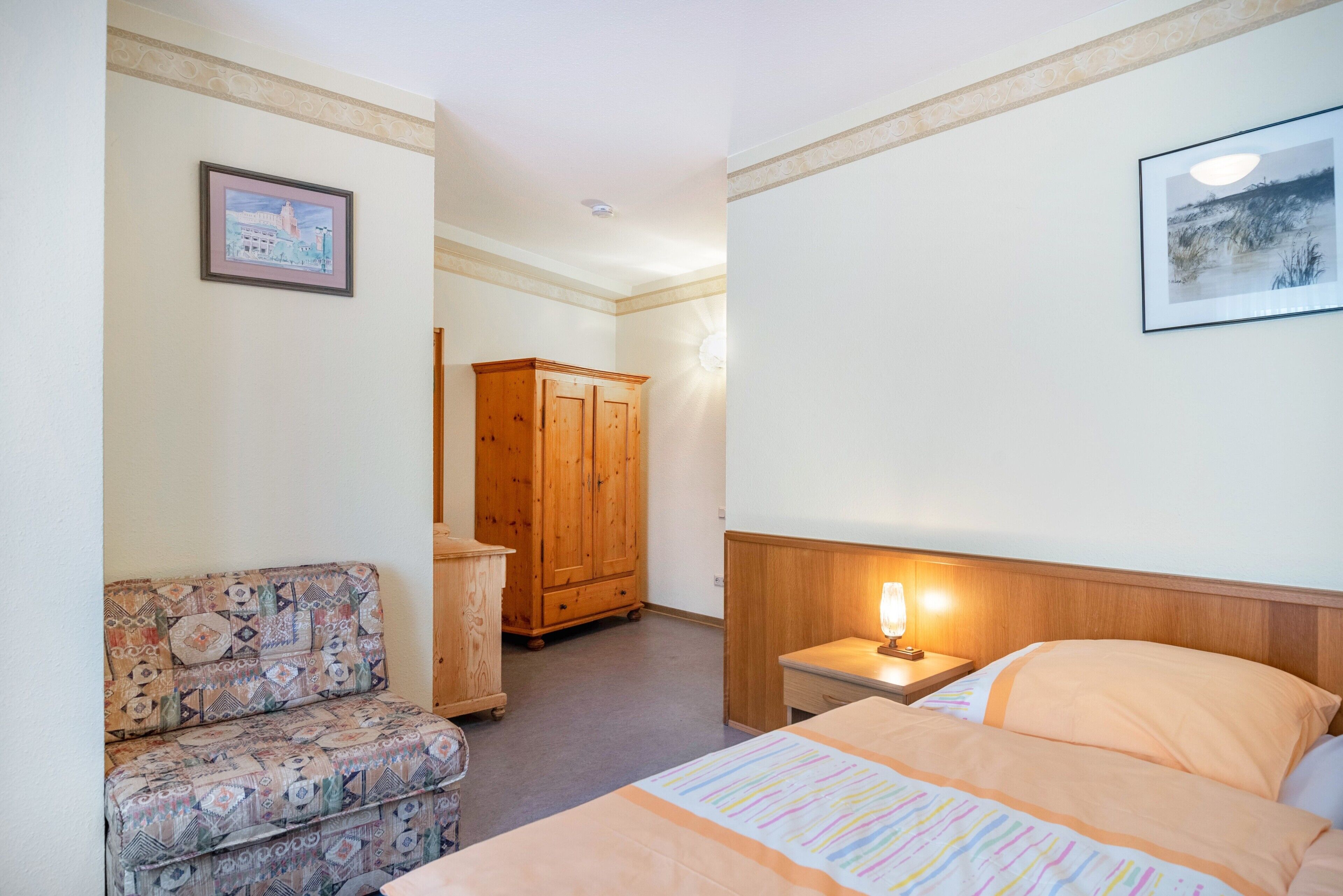 A hotel room with a bed, a nightstand with a lamp, a wooden wardrobe, a patterned armchair, and two framed pictures on the walls.