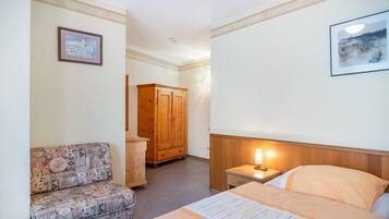 A hotel room with a bed, a nightstand with a lamp, a wooden wardrobe, a patterned armchair, and two framed pictures on the walls.