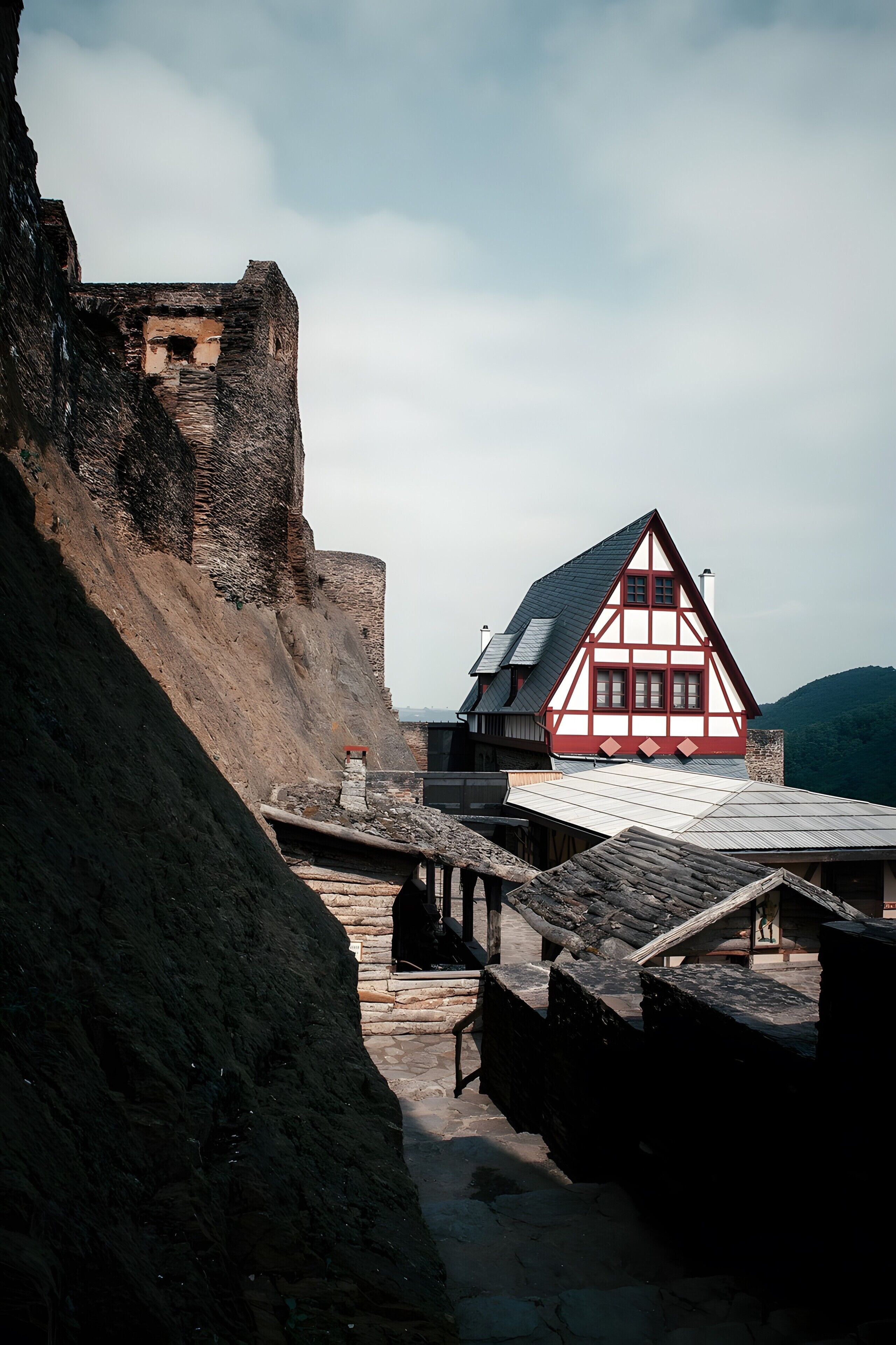 A traditional half-timbered house with a stone wall and a steep roof, located near a rocky cliff.