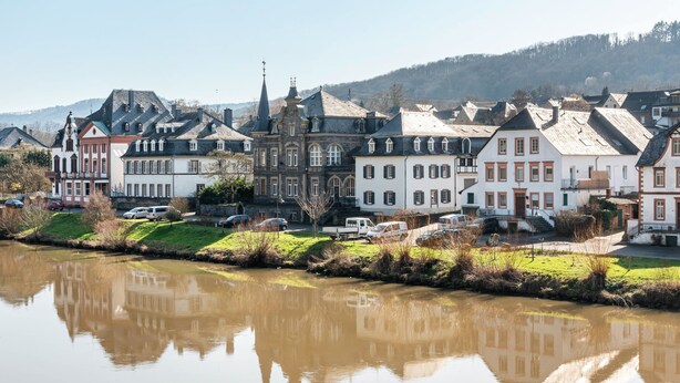 Een pittoresk stadje aan de rivier, met historische gebouwen, een kerktoren en een rustig waterlichaam dat de architectuur weerspiegelt.