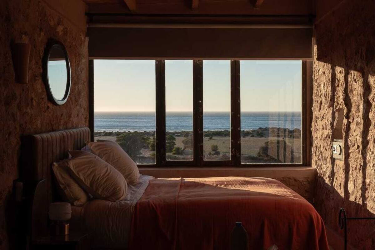 A bedroom with a large window overlooking the sea, a bed with pillows, and a bedside table with a lamp.