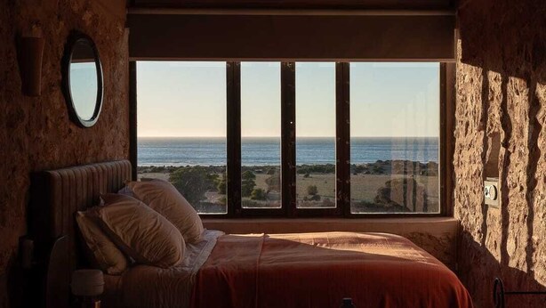 Un dormitorio con un ventanal grande con vistas al mar, una cama con almohadas y una mesita de noche con una lámpara.