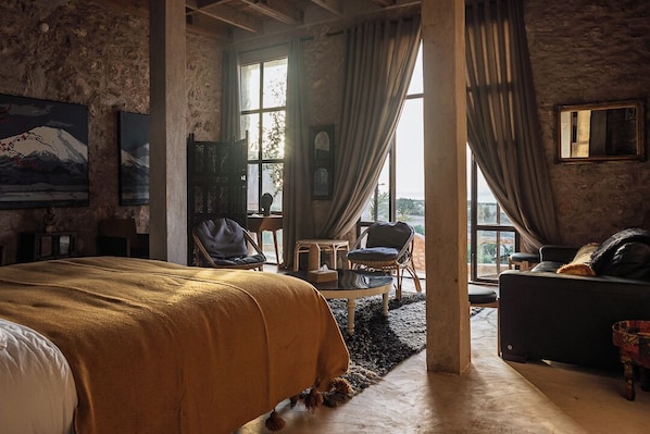 A spacious room with a large bed, a sofa, and a chair. There is a window with curtains, a painting on the wall, and a view outside.