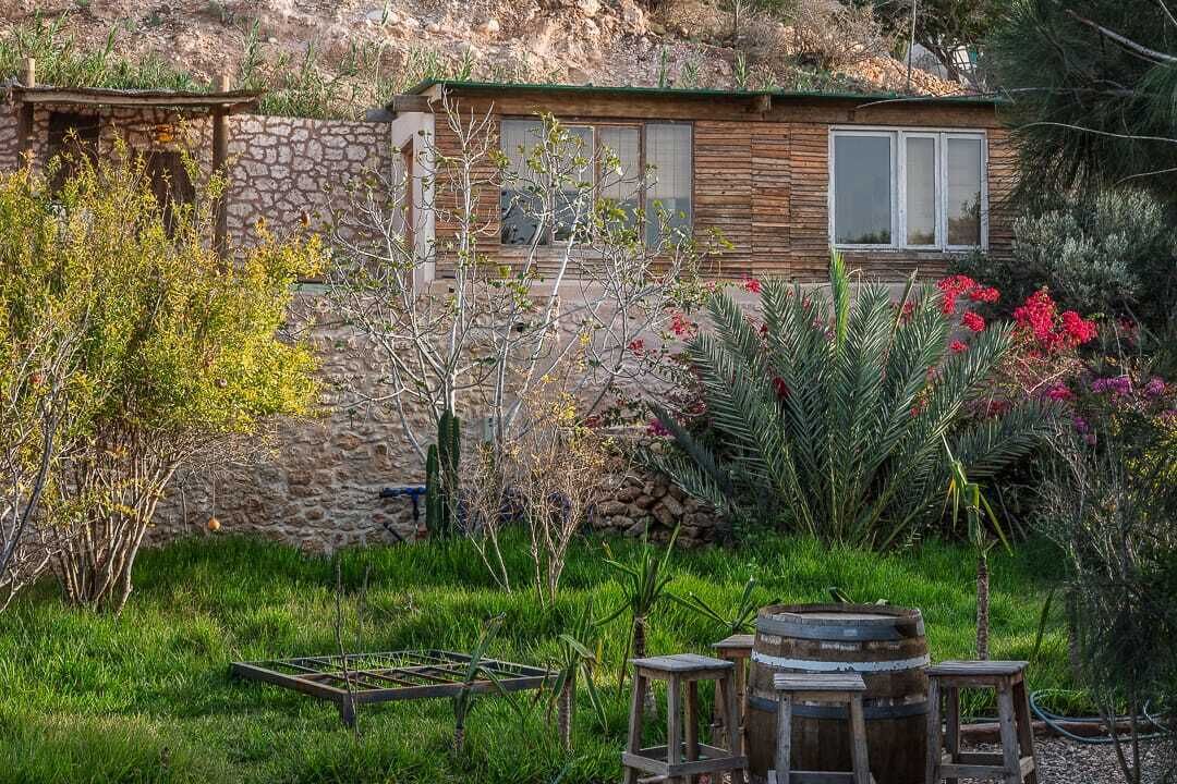 Uma casa rústica com parede de pedra, janelas de madeira e um jardim com diversas plantas e um barril de madeira.