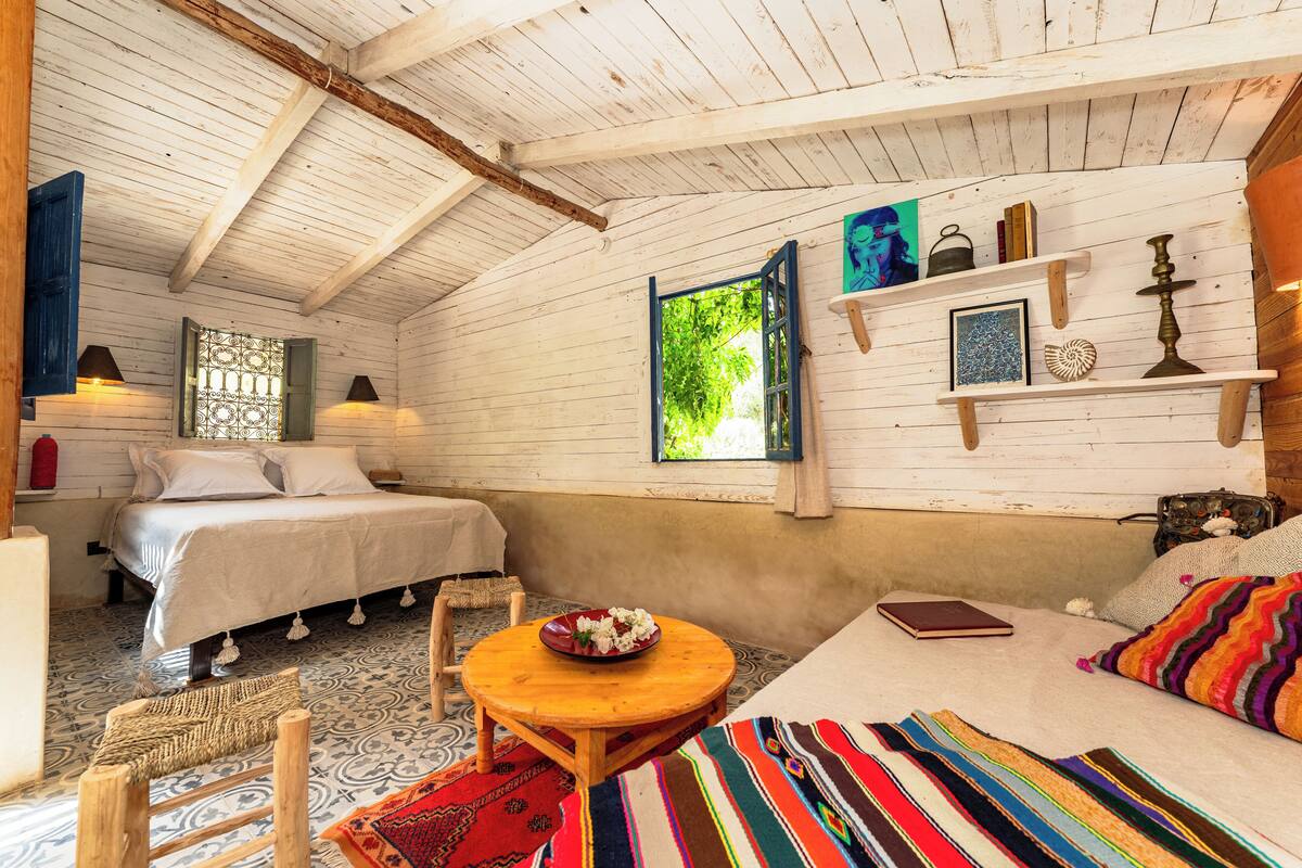 A room with two beds, a small table with a bowl, a sofa, and a window with a view of greenery.