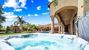 A hot tub area with a pool, palm trees, and a building with arches. - Luxury 6BR Mesa Estate | Pool, Water Slide, Pickleball, Theater (Mesa)