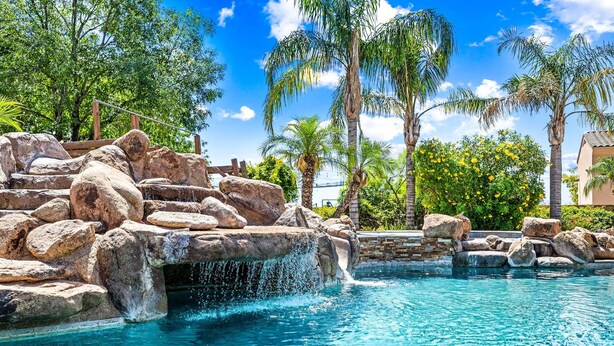 A pool with a waterfall feature surrounded by rocks and palm trees. - Luxury 6BR Mesa Estate | Pool, Water Slide, Pickleball, Theater (Mesa)